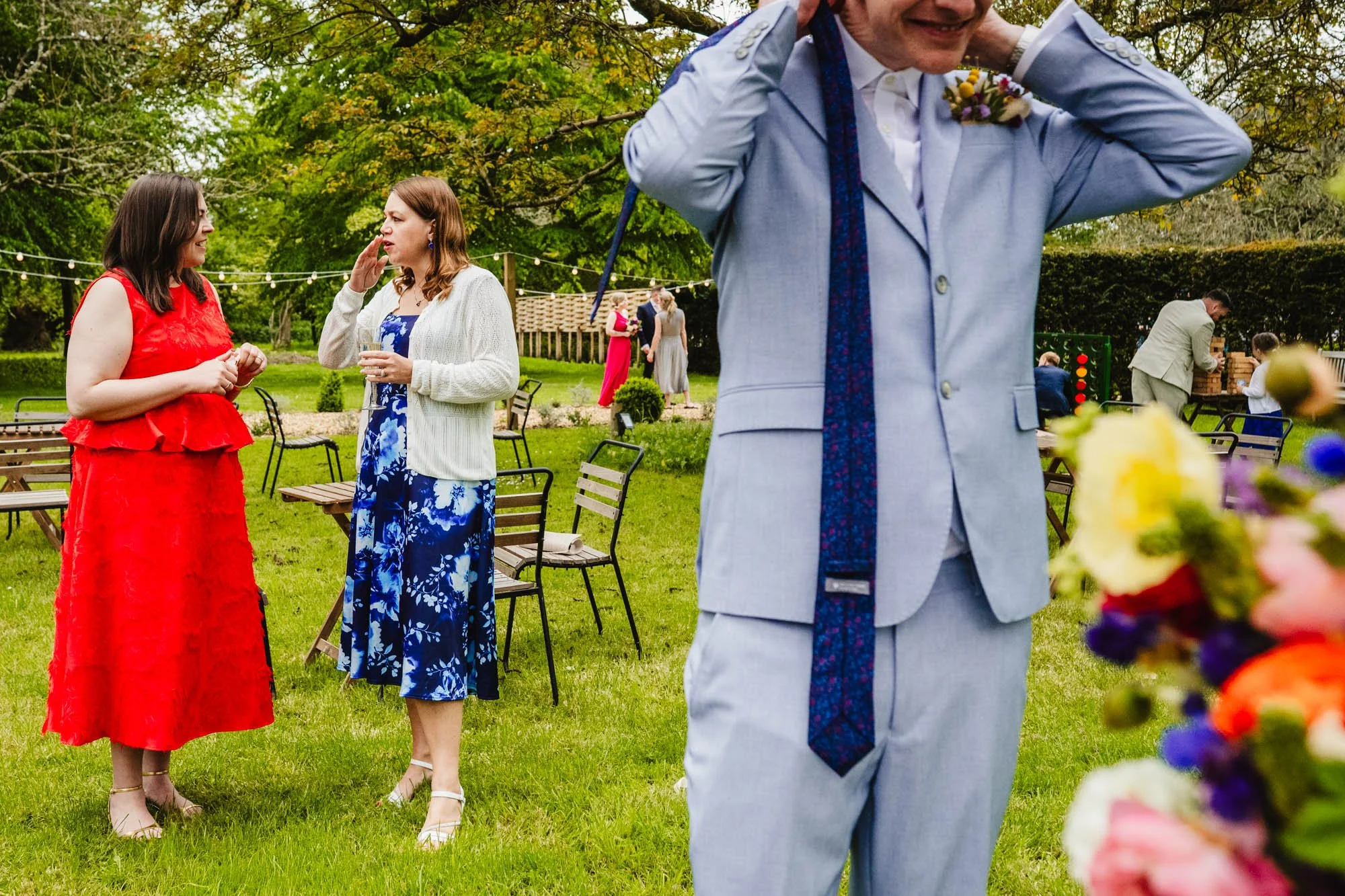 A candid, layered photograph of an outdoor wedding reception with a man putting his tie on in the foreground and clusters of people in the background