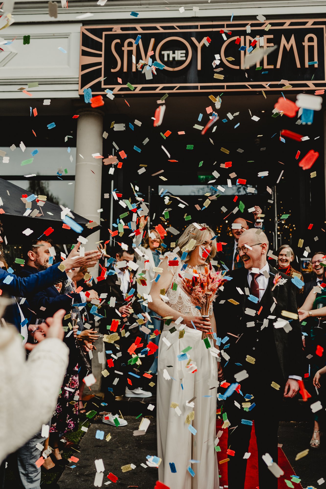 Photograph of a couple standing on a red carpet outside the Savoy Cinema in Heaton Moor while their guests throw movie stubs as confetti just after their wedding ceremony