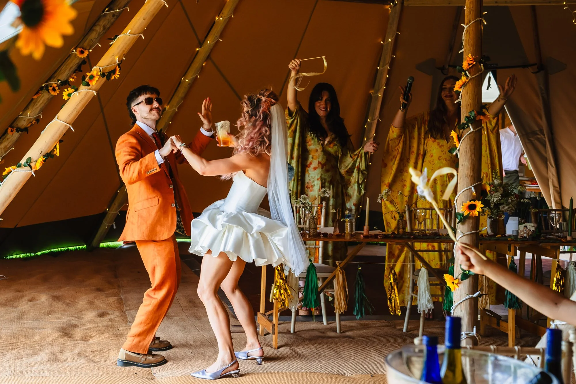 Photograph of a bride and groom making an entrance to their wedding breakfast in a tipi at Duddon Mill Farm. They are dancing while guests wave ribbons in the air.