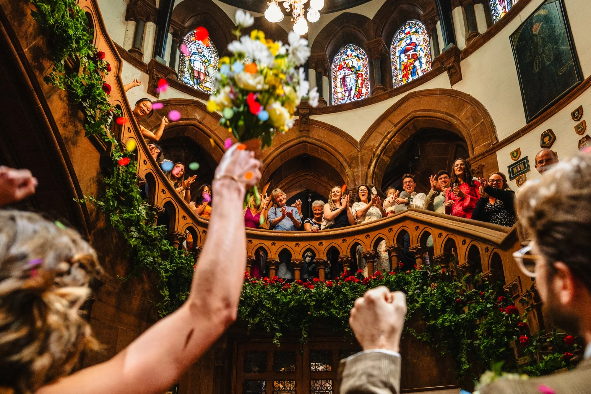 photograph of a group of wedding guests on the inner stairs at Chester town hall. They are all clapping and cheering at the bride and groom below, with some colourful confetti still floating in the air.