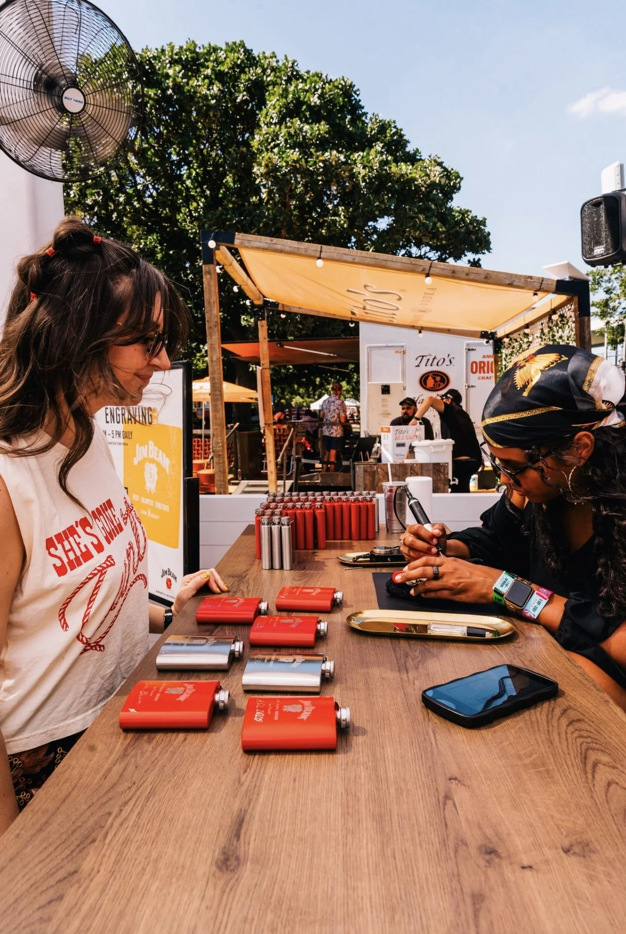 Two women are sitting at a wooden outdoor table with red and silver flameless heat packs; one woman is signing an item while the other is smiling and watching. Behind them are food stalls and people, with trees and a blue sky overhead.