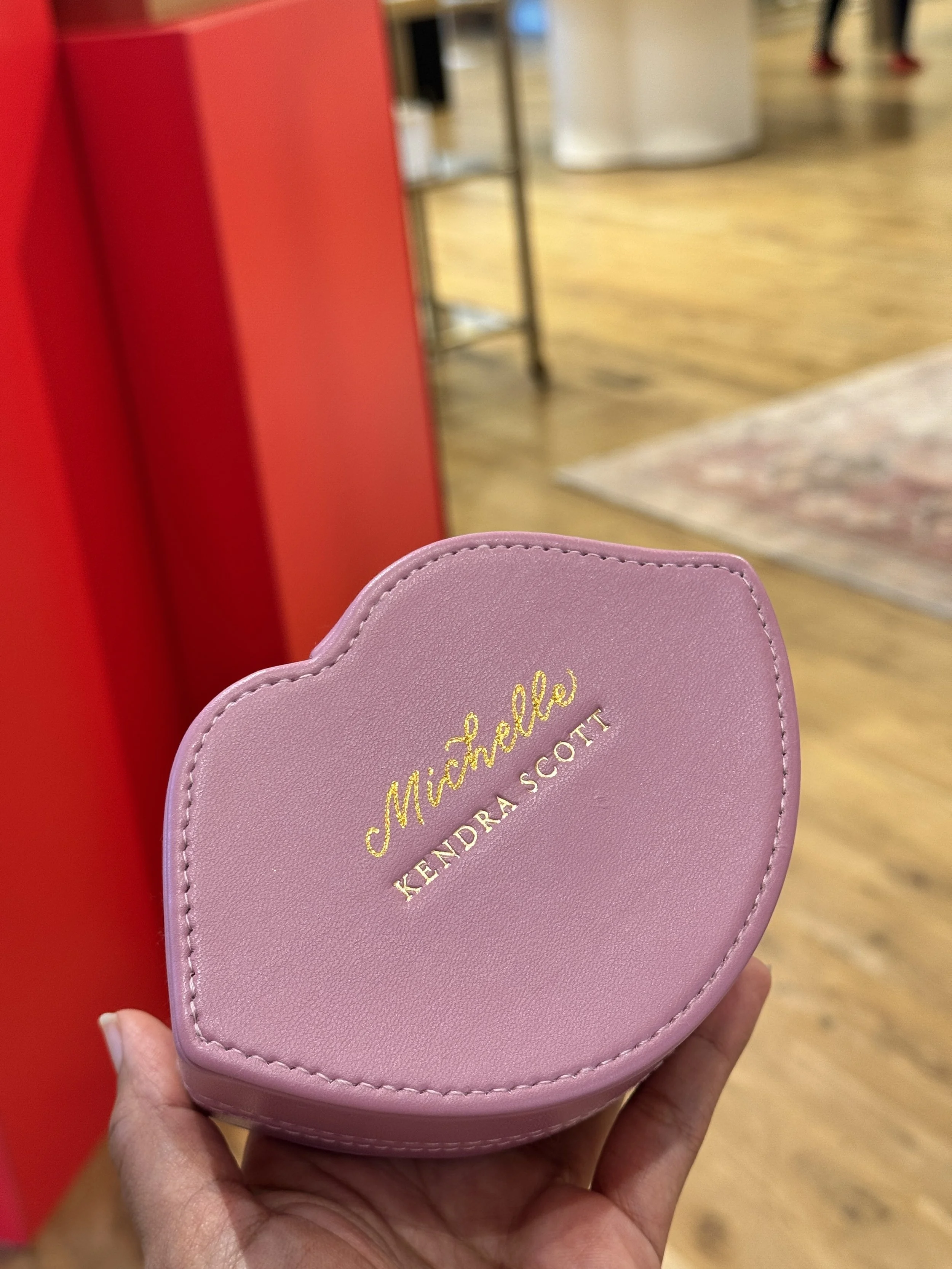 A pink Michael Kors small handbag with gold lettering, held in a person's hand in a store with wooden flooring and red and white displays.