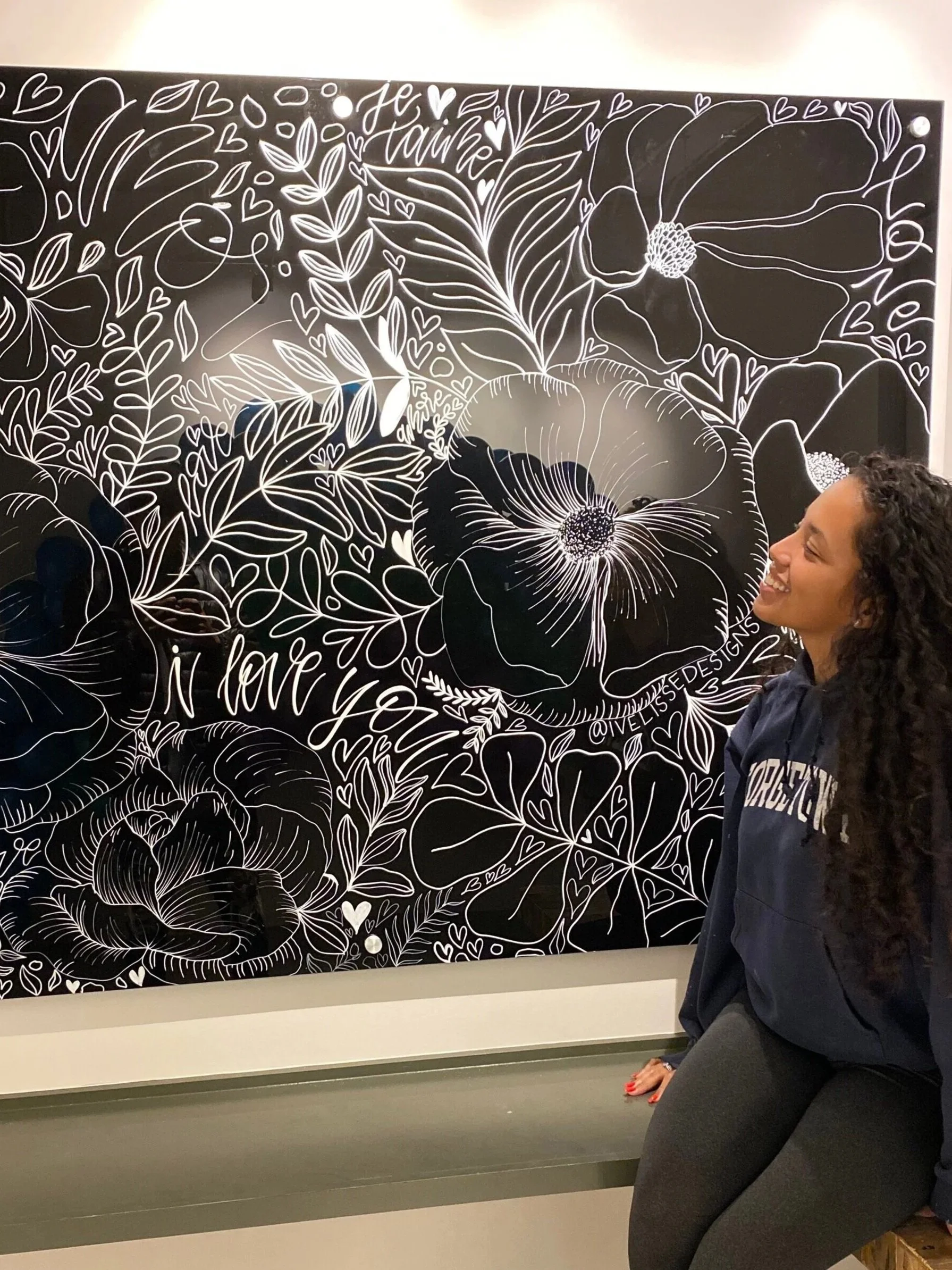 Close-up of a black wall with white floral and heart line art drawings, including large flowers, leaves, and words like 'love' and 'faith' in cursive, with a smiling woman sitting in front of it.