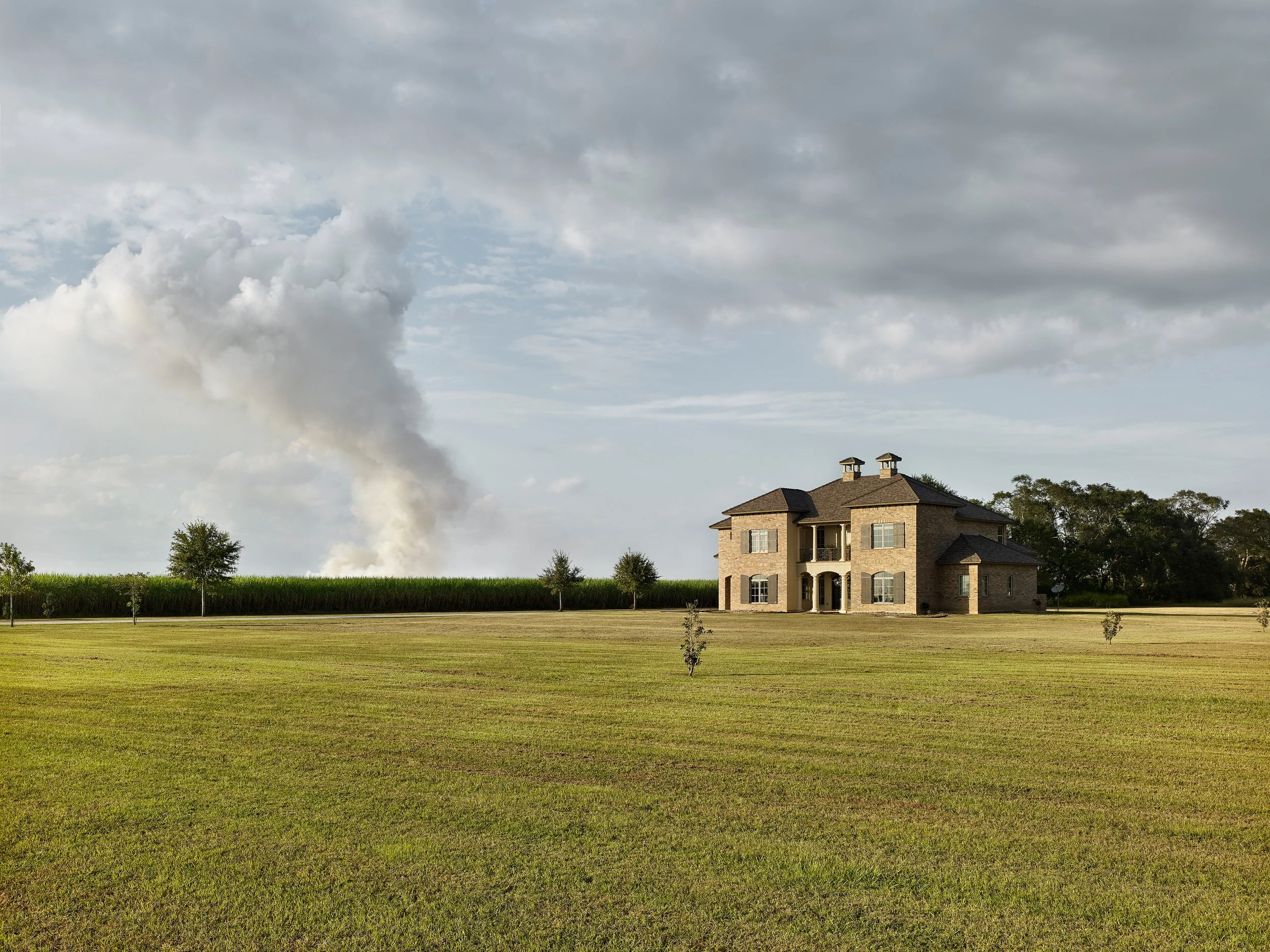 Large brick house beside a cornfield and expansive grassy lawn under dramatic cloudy skies