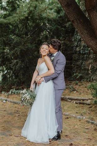 A bride and groom embracing outdoors in a wooded area, with the groom kissing the bride on her cheek, both smiling.