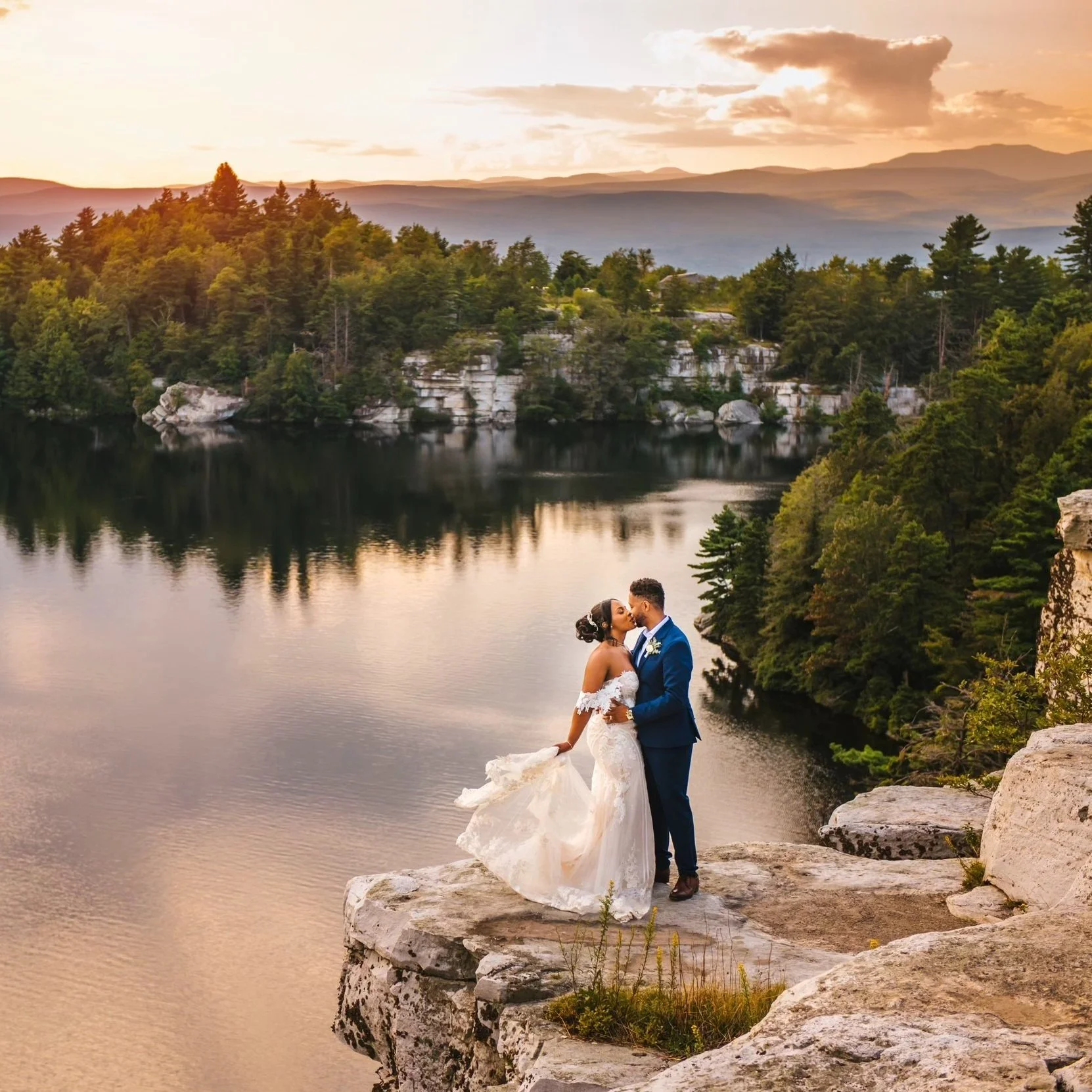 A bride and groom standing on a rocky ledge by a calm lake at sunset, kissing with trees and mountains in the background.