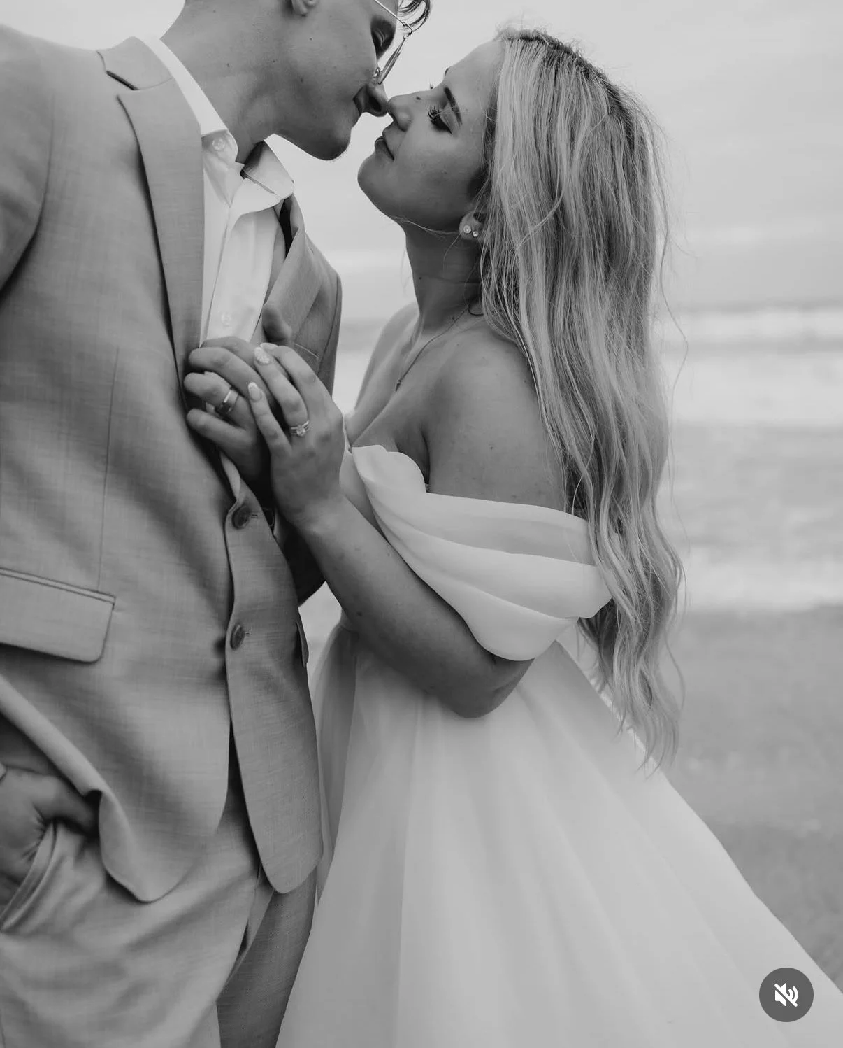 A black and white photo of a couple on the beach, close-up, touching noses, with the woman holding the man's suit lapel and both smiling softly.