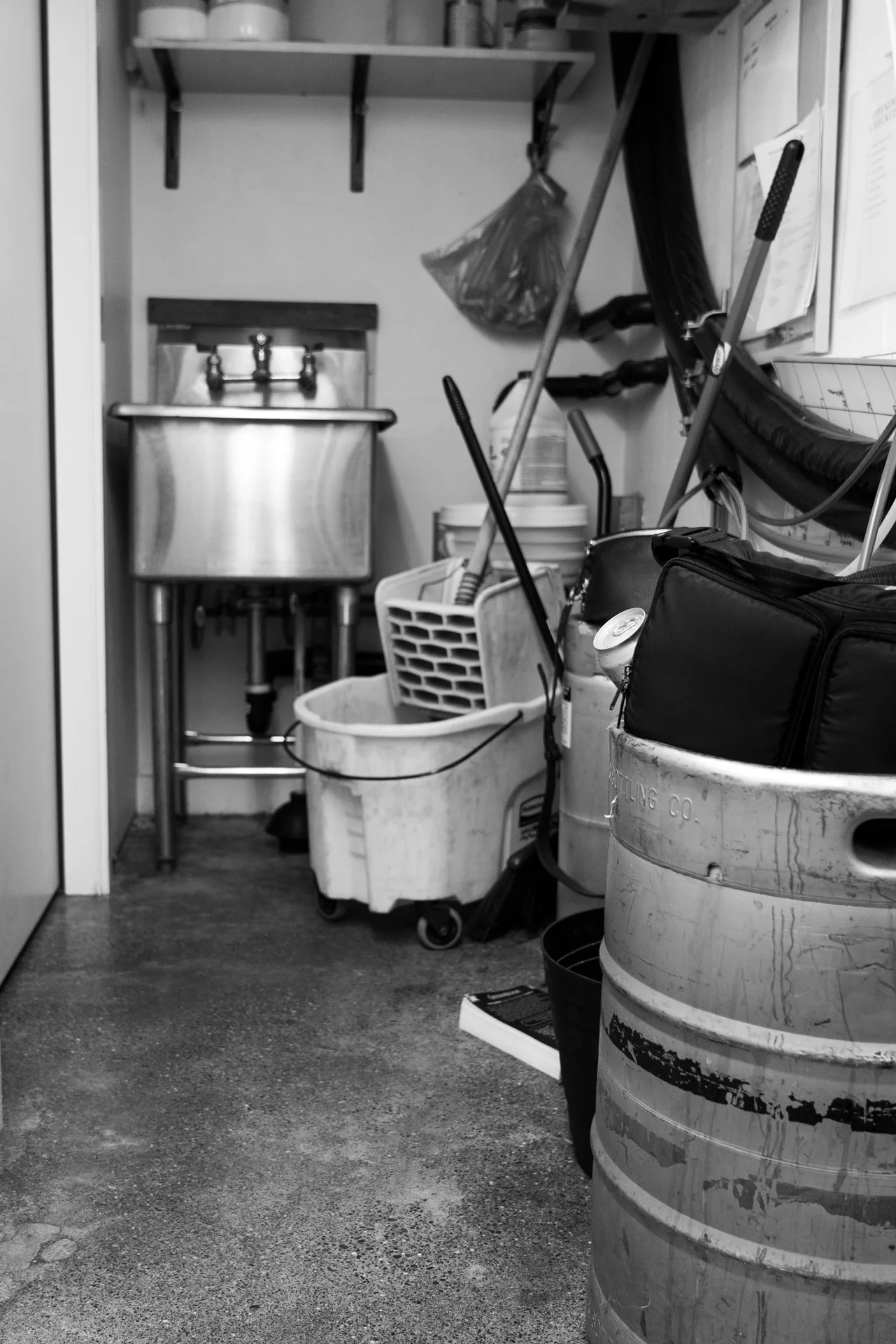 A black and white photo of a cluttered utility or maintenance closet with cleaning supplies, buckets, a mop, and a sink.
