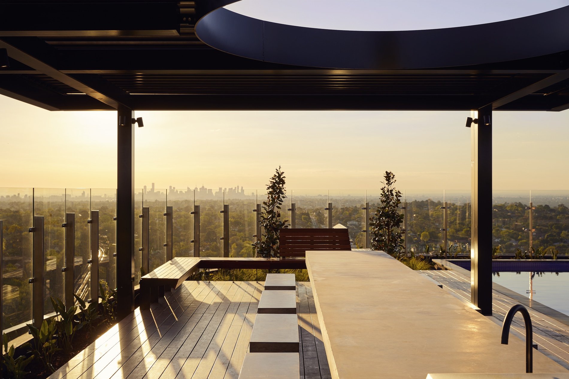 Rooftop lounge area with a wooden bench, table, and plants, overlooking a city skyline at sunset.