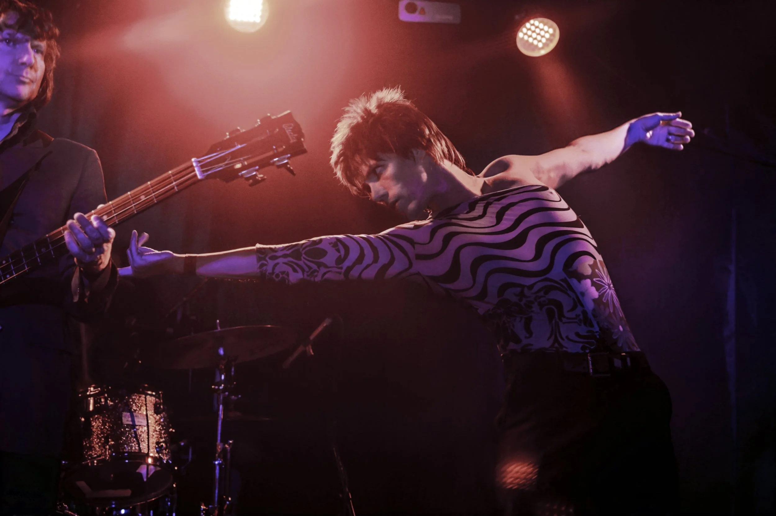 Musicians performing on stage with red and purple lighting, one playing guitar and the other dancing or singing