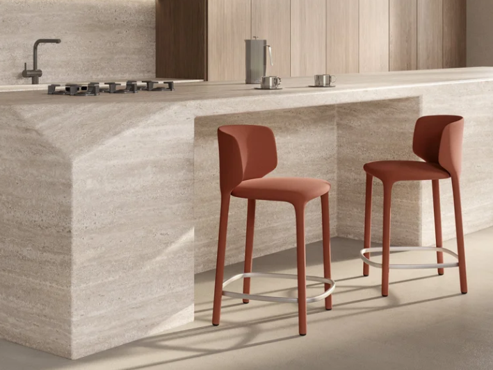 Modern Pat stools featuring terracotta-red upholstered seats, low rounded backrests, and metal footrests, styled along a light travertine kitchen island.