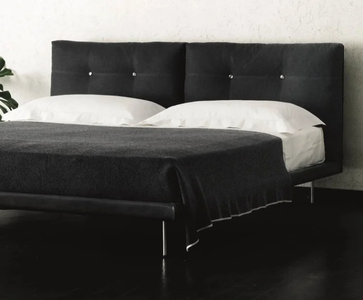 Rod bed that features a dark upholstered tufted headboard, slender metallic legs, crisp white pillows, and a charcoal gray blanket, styled against a textured light wall.
