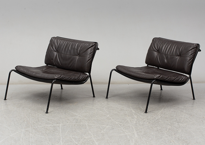 Pair of Frog lounge chairs with tufted dark brown leather cushions on thin black metal frames with splayed legs, photographed on a light concrete floor.