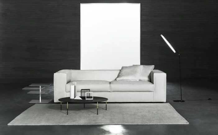 : Neowall sofa in white upholstery with clean-lined modern design, positioned against a dark textured wall with bright white panel, accompanied by a black round coffee table, floor lamp,
