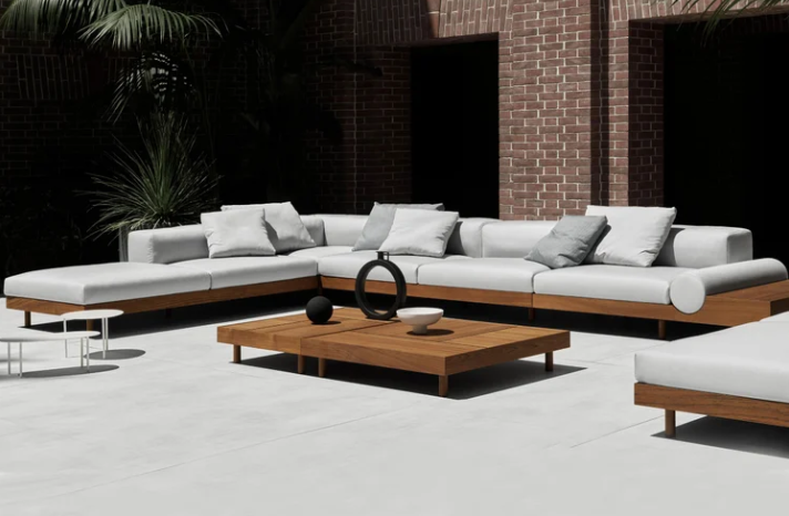 Kasbah outdoor collection featuring a large wooden sectional sofa with plush light gray cushions, paired with matching low wooden coffee tables on a patio against a brick wall background.