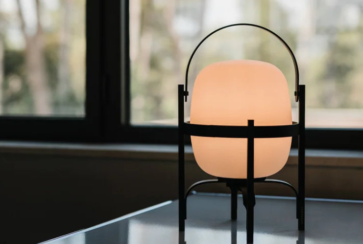 Cesta table lamp featuring an opal glass shade encased in a dark lantern-style frame with an arched top handle, glowing brightly next to a large window.