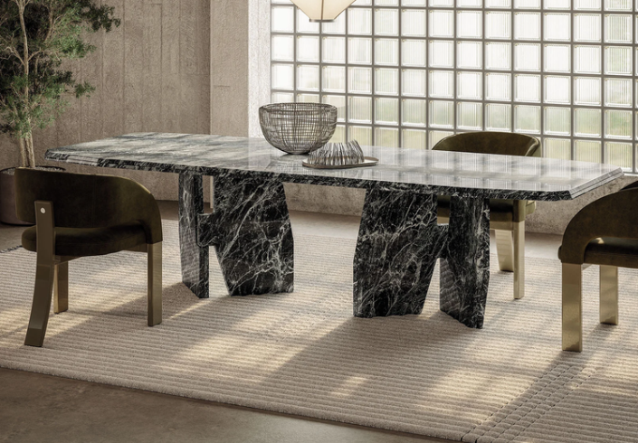 Fosca dining chairs in olive green velvet with brass legs surrounding a sculptural black marble dining table with organic-shaped pedestal bases, set on a textured beige rug against a glass block wall.