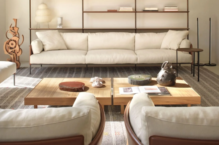 Greene sofa featuring light cream fabric upholstery, plush cushions, and slender dark metal legs, styled with low wooden coffee tables and warm interior decor.
