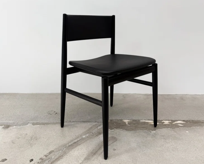 Modern Neve chair featuring a sleek black wooden frame with a gently curved backrest and leather upholstered seat, set on a plain concrete floor against a white wall.