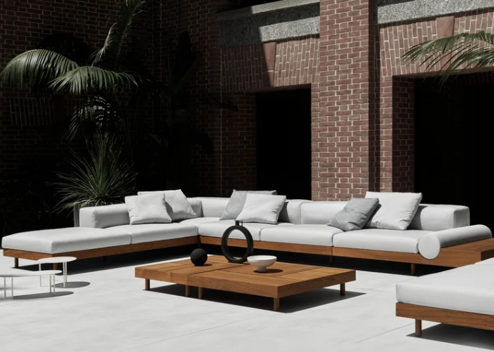The Kasbah outdoor sectional sofa features thick white cushions on a natural wood base, arranged in a U-shape around a matching wooden coffee table in a modern patio courtyard with brick walls and palm trees.