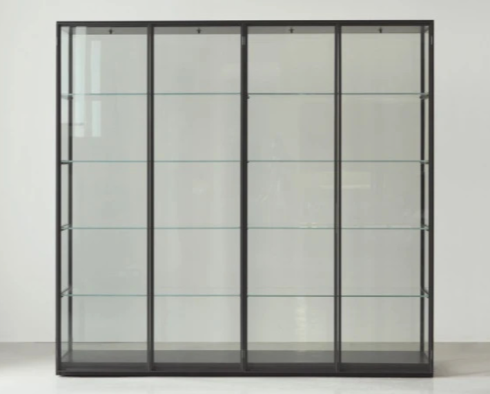 Ex-Libris bookshelf featuring a sleek dark metal frame enclosing four glass cabinet sections with transparent glass shelves, set against a plain white wall.