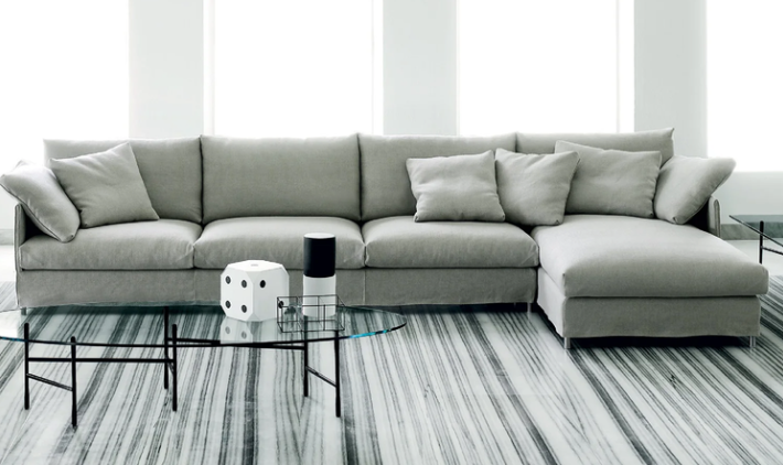 Chemise sectional sofa upholstered in light grey fabric with plush cushions and throw pillows, featuring an attached chaise lounge and styled over a striped rug.