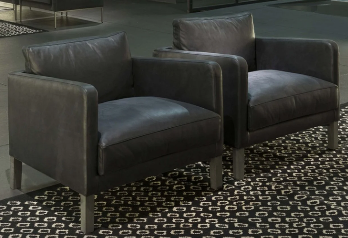 Dumas armchair featuring dark charcoal grey leather upholstery with deep seat cushions and sturdy square legs, displayed as a pair on a black and white patterned rug.