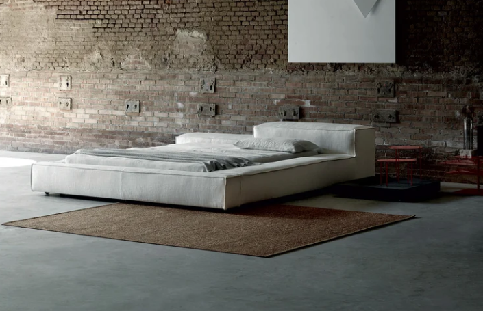 Extrasoft bed featuring a low-profile fully upholstered platform frame in off-white fabric with plush headboard cushions, styled on a brown area rug.