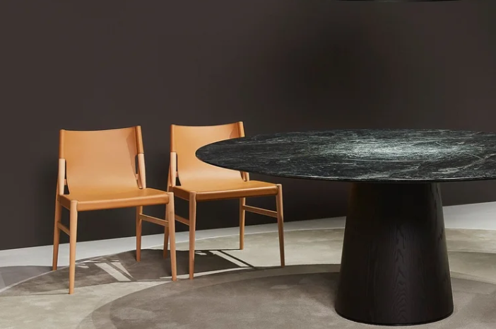 Two Voyage dining chairs in tan leather with light wood frames positioned beside a round black marble dining table with a dark pedestal base against a charcoal wall.