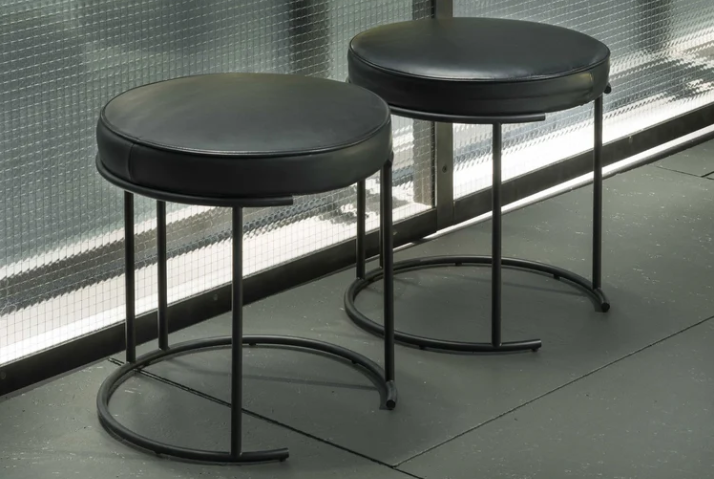 Nina stools featuring round black leather cushioned seats supported by minimalist black tubular metal frames with curved bases, shown as a pair against a glass-paneled wall.