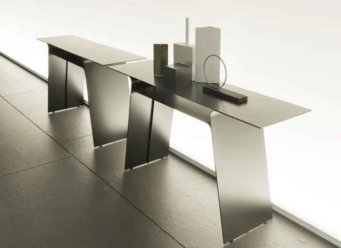Sleek Inari console tables with a minimalist metallic design, thin tabletop surface, and angled sheet-metal legs styled with geometric decor pieces against a bright wall.