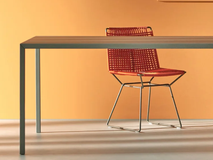 Tense outdoor dining chair with red woven mesh back and seat on a chrome metal frame, positioned at a minimalist table with thin metal legs against a warm orange wall.