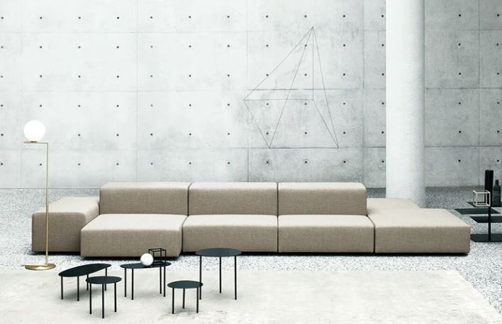 Extra Wall modular sofa featuring a low-profile geometric design in neutral beige fabric, styled with small black accent tables and a slender floor lamp against a concrete wall.
