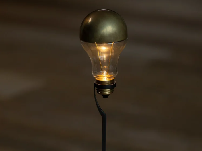 Trenta table lamp featuring an exposed glass light bulb with a metallic brass dome cap, glowing warmly atop a slender black metal stand against a soft, dark background.