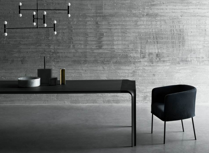 Brasilia dining table in black with distinctive curved leg detail, paired with a black upholstered chair and geometric chandelier against a textured gray concrete wall.