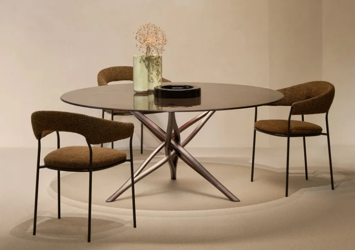 SK dining table with a round glass top supported by a central, multi-legged metallic base with intertwining branches, surrounded by textured brown dining chairs.