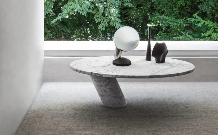 Eccentrico table featuring an elliptical white marble tabletop supported by an asymmetrical, slanted cylindrical base, styled with a spherical table lamp and decor accents in front of a floor-to-ceiling window.