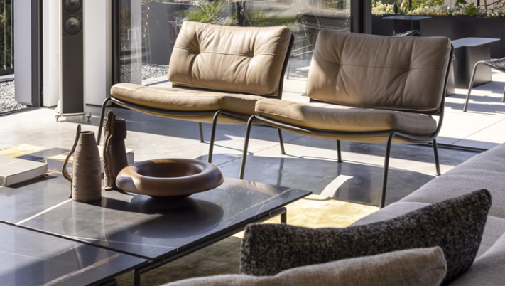 Frog lounge chairs featuring low-slung dark metal frames with tufted tan leather seat and back cushions, shown as a pair in a sunlit living space.