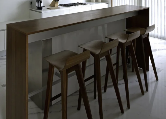 Morph stools featuring sculpted solid wood seats with low back lips and slender angled legs, lined up along a dark wooden counter in a contemporary kitchen setting.