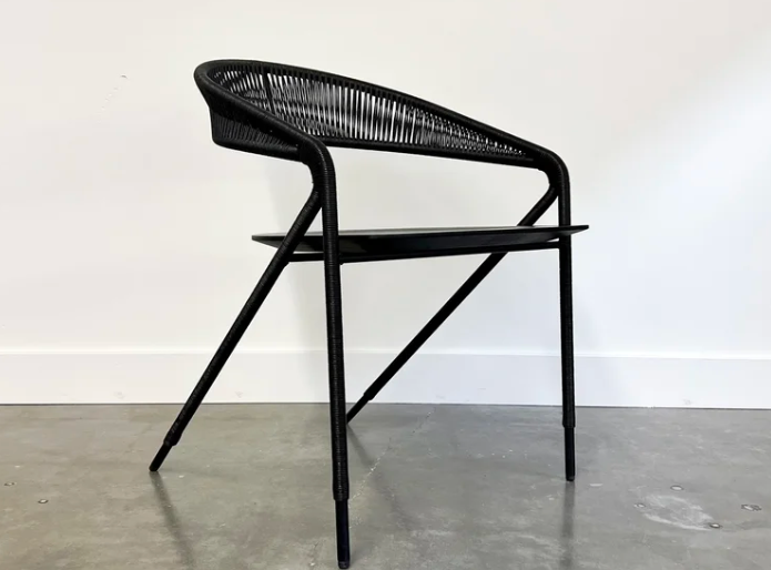 Georges dining chair in black featuring a sculptural cord-wrapped backrest with vertical weaving, flat seat, and angled tapered legs, photographed against a white wall on a concrete floor.