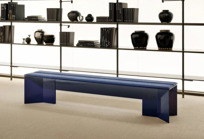 Origata bench featuring a vibrant blue geometric sheet-metal frame topped with split cushioned seating, positioned in front of a dark floor-to-ceiling shelving unit.