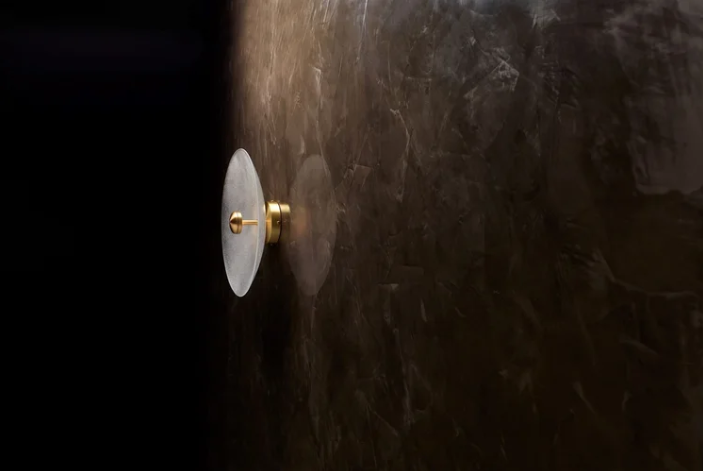 Cielo wall lamp featuring a circular frosted glass disc shade with brass mounting hardware, casting a soft glow against a dark textured marble wall.