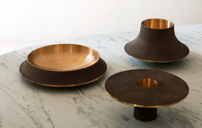 Modern Cap53 decorative objects featuring dark brown textured exterior finishes with polished bronze interiors, arranged in bowl and pedestal forms on a white marble surface.