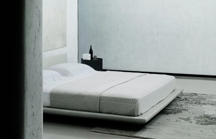 Chemise low-profile platform bed with white upholstered frame and bedding, featuring a minimalist floating design with textured throw blanket, set against light walls with a black cylindrical side table.