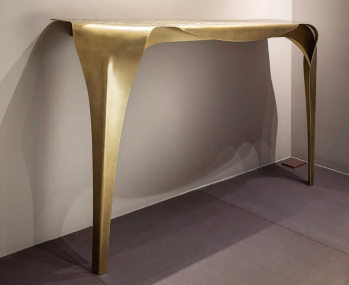 Convivium console table featuring a fluid, sculptural brushed brass frame with draped, organic legs, leaning against a neutral wall in a minimalist interior.