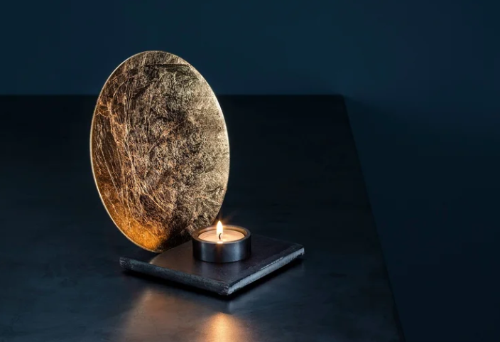Sculptural Luna objet candleholder featuring a textured gold leaf disc that reflects the warm glow of a small lit tealight on a dark square base.