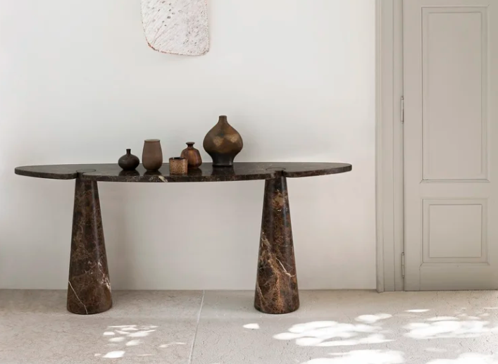 Sculptural Eros console table in dark marbled stone featuring two conical legs supporting an elongated, rounded tabletop with decorative ceramic vases against a white wall.