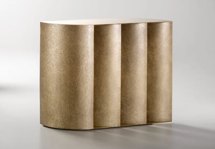 Sinestesia console table that has a textured brass finish and a sculptural base with vertical curved panel columns, isolated against a light gray background.