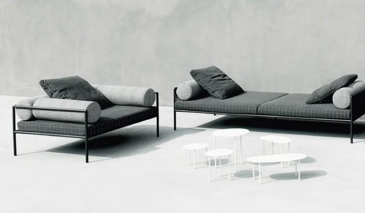 Agra outdoor sofa setup featuring sleek black metal frames, dark patterned upholstery, and light bolster cushions, styled alongside low white side tables against a concrete wall.