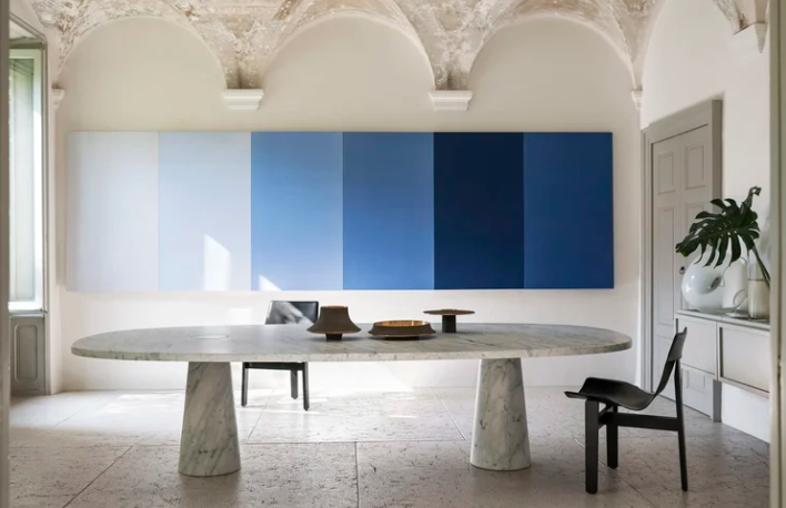 Modern Eros dining table with an oval marble top supported by two thick conical marble pedestals, styled with dark chairs in an architectural space with vaulted ceilings.